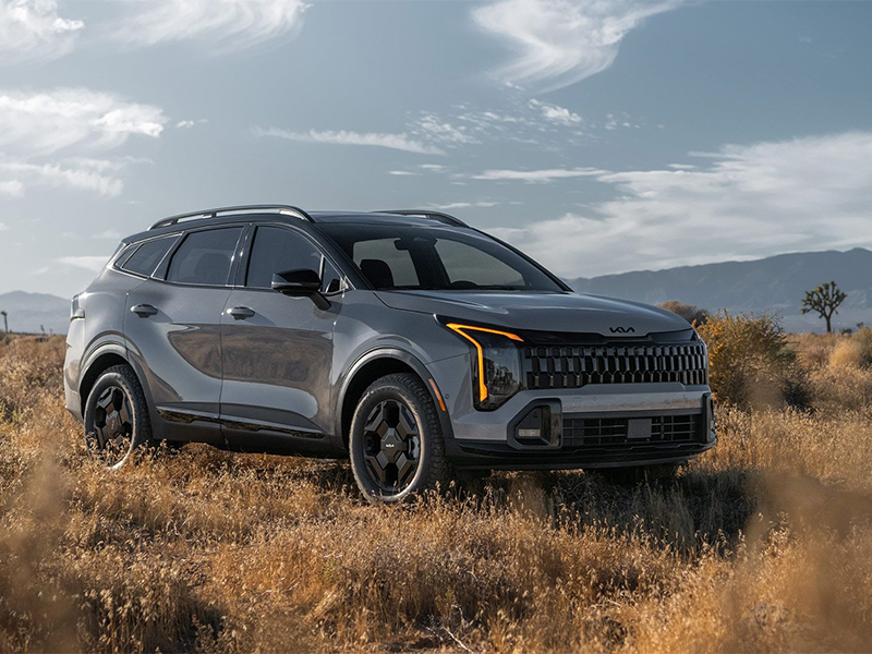 A gray 2026 Kia Sportage SUV parked in a dry, grassy landscape with mountains in the background, showcasing its bold front grille and modern LED lighting design.