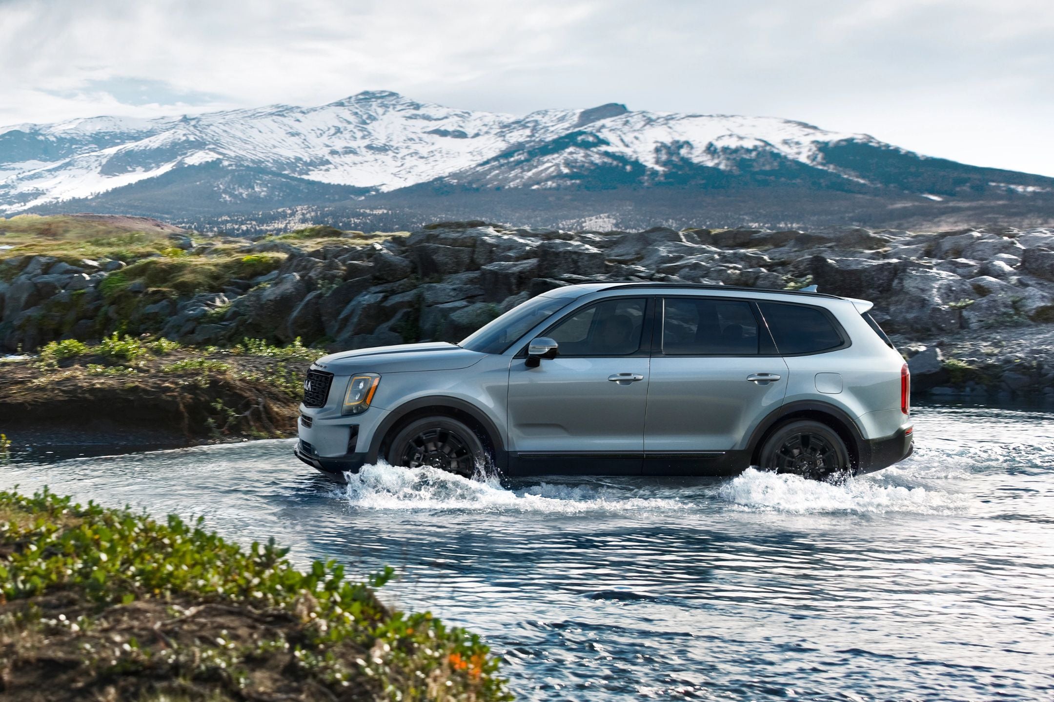 A silver SUV drives through a shallow stream with snow-covered mountains in the background.